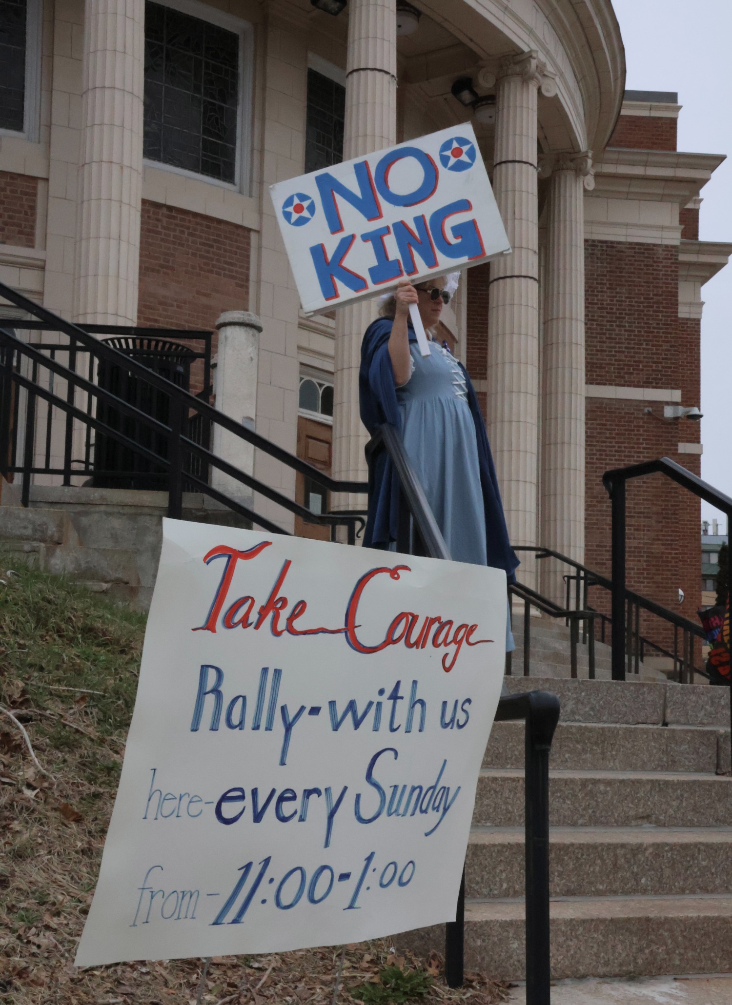 A protester holds a “No Kings” sign on the steps of Hamden Town Hall, standing behind a posted notice inviting community members to join weekly demonstrations at the same location every Sunday from 11 a.m. to 1 p.m. Hannah Freshman/HQNN