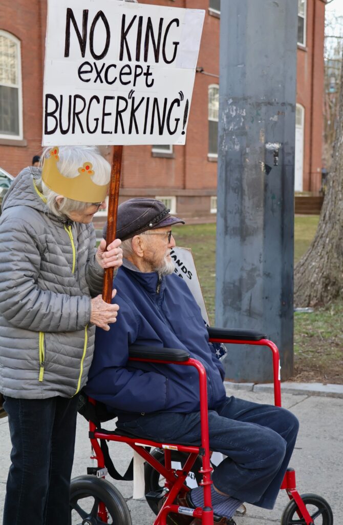 At the end of the route, two people watch the march, with one seated in a wheelchair and the other holding a sign reading “No king except Burger King.” (Hannah Freshman/HQNN)