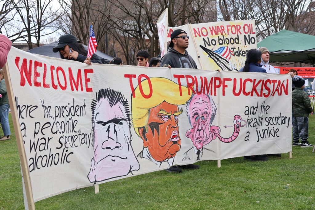 A large hand-painted sign featuring portraits of Donald Trump and Robert F. Kennedy Jr. and the message “Welcome to Trumpfuckistan” is displayed. Hannah Freshman/HQNN