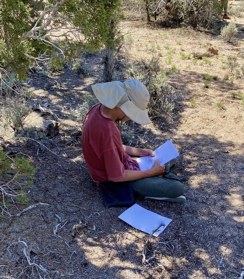 A photo of Eric from behind sitting while reading letters from his parents while at Evoke Therapy Program's Wilderness Therapy Program in Santa Clara, Utah. He is wearing a sun hat, a t-shirt and pants. 
