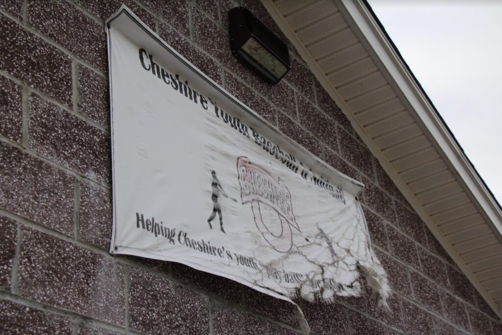 A faded and tattered Cheshire Youth Baseball and Softball sign overlooks the empty recreational fields at Bartlem Park in Cheshire. 
Photo Credit: Lo Yarnall